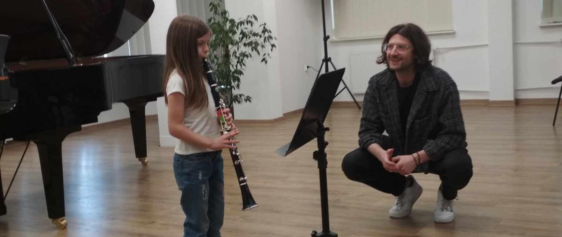 Warsztaty klarnetowe. Mała dziewczynka w białej koszulce i granatowych spodniach gra na klarnecie. Przed nią pulpit na nuty. Obok niej kuca mężczyzna i przysłuchuje się jej grze. W tle stoi czarny fortepian.