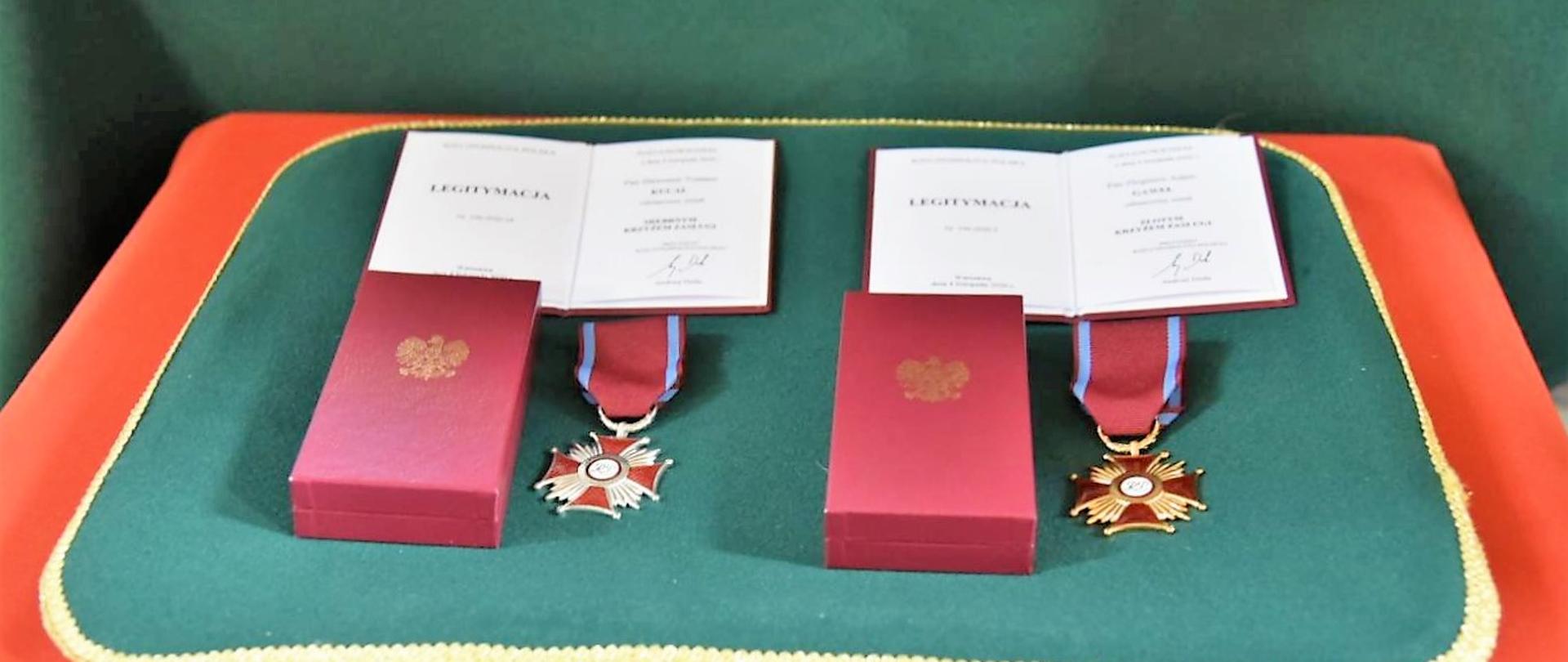 Na zielonej, ozdobnej poduszce leżą 2 odznaczenia, 2 pudełka na odznaczenia i dwie legitymacje dla odznaczonych 
