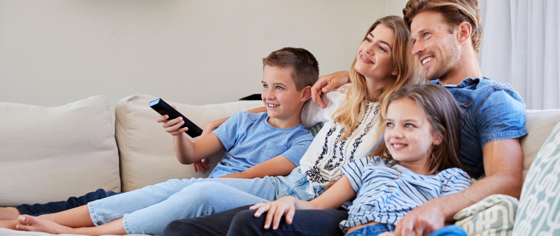 Uśmiechnięta rodzina z dwójką kilkuletnich dzieci na kanapie. Chłopiec trzyma w dłoni telewizyjnego pilota.