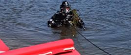 Ratownik w czarnym kombinezonie do nurkowania wyłania się z wody przy brzegu zalewu, trzymając w rękach rośliny wodne. Na pierwszym planie czerwony ponton ratowniczy z napisem „OSP Pakosław”.