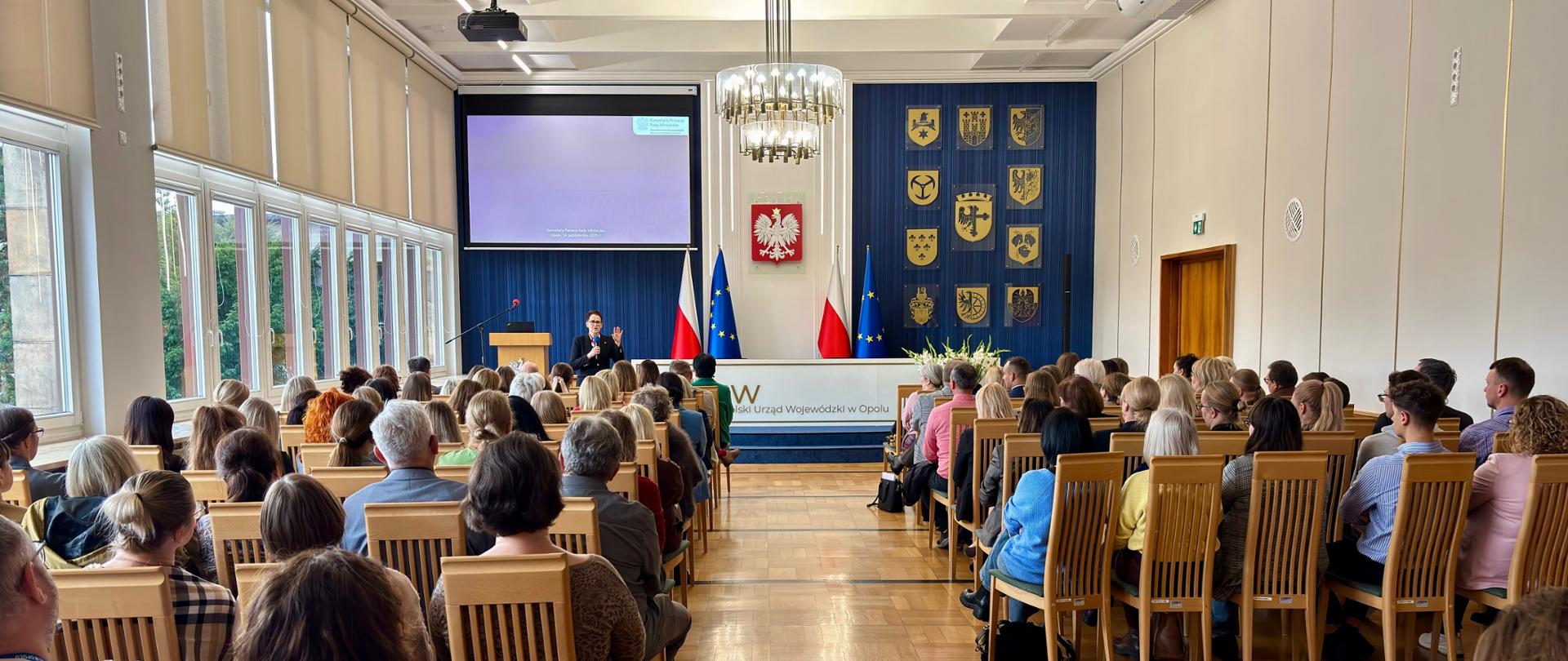 Szefowa służby cywilnej przemawia w dużej jasnej auli w urzędzie. Na środku ściany godło Polski.