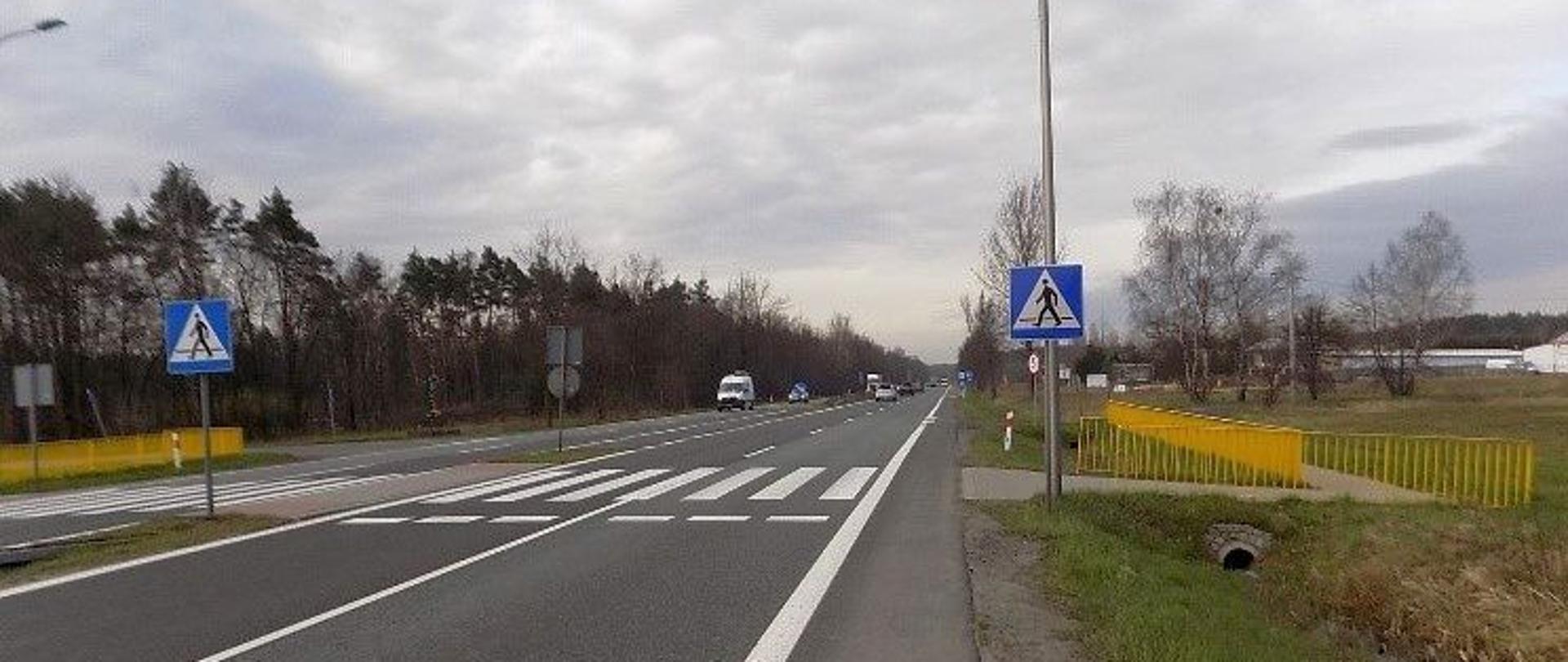 Fotografia przejścia dla pieszych na DK86 w Kuźnicy Podleśnej. Przejście zlokalizowane w terenie mało zurbanizowanym. Dojście do przejścia wygrodzone barierami ochronnymi w żółtym kolorze. Na drodze dwujezdniowej widoczne poruszające się po drodze pojazdy. 