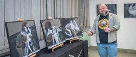 Zdjęcie przedstawia słuchacza pozującego na tle swojej wystawy fotograficznej. Obok niego na stole przykrytym czarnym obrusem też stoją zdjęcia na drewnianych sztalugach. 