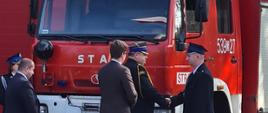 Przed samochodem gaśniczym stoi mężczyzna obok strażacy podają sobie ręce.