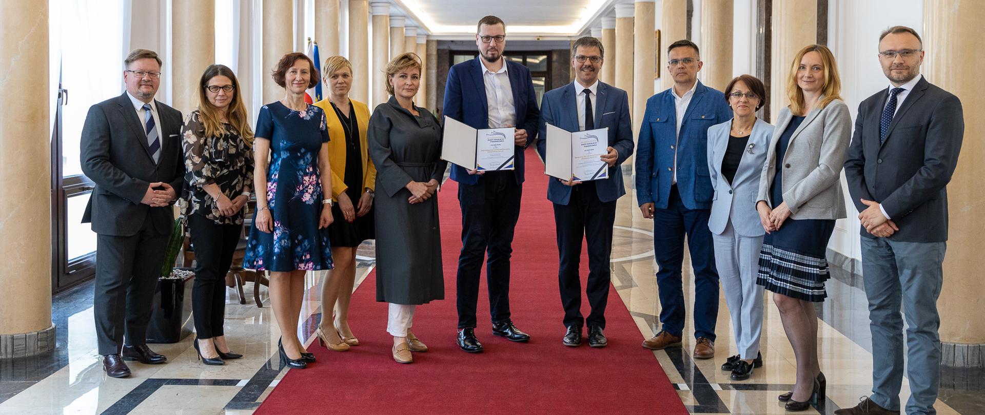 Uczestnicy uroczystości wręczenia wyróżnień Rady Edukacji Finansowej za 2021 rok, na korytarzu w gmachu MF (czerwony dywan, kolumny)