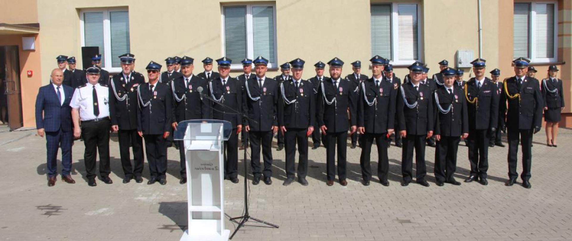 Strażacy pozujący do pamiątkowego zdjęcia. Poza druhami z Ochotniczej Straży Pożarnej w Wiśniewie na zdjęciu znajduje się również Komendant Powiatowy Państwowej Straży Pożarnej w Zambrowie młodszy brygadier Andrzej Kalinowski oraz Wójt Gminy Zambrów Pan Jarosław Kos. Strażacy ubrani w mundury wyjściowe ze sznurem galowym. Pamiątkowe zdjęcie wykonano w słoneczny dzień.