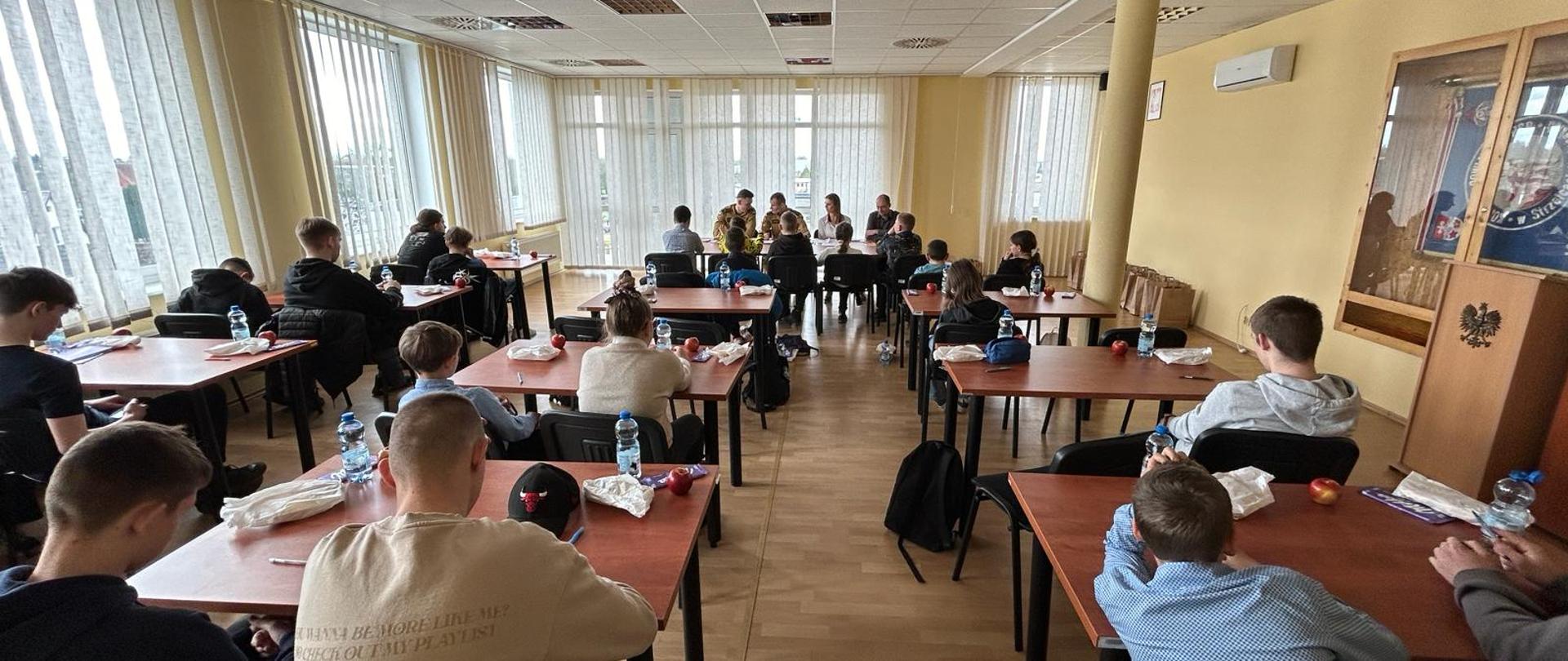 Fotografia przedstawia ujęcie pomieszczenia – świetlicy w Komendzie Powiatowej Państwowej Straży Pożarnej w Strzelcach Krajeńskich w trakcie trwania eliminacji powiatowych Ogólnopolskiego Turnieju Wiedzy Pożarniczej, w głębi pomieszczenia przy stole członkowie komisji konkursowej przeprowadzają część ustną eliminacji - odpytują uczestników.