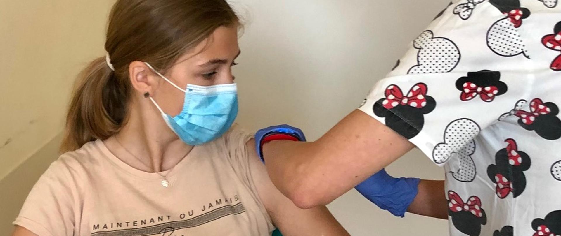 Dziewczynka p[przyjmuje szczepienie z rąk pielęgniarki 