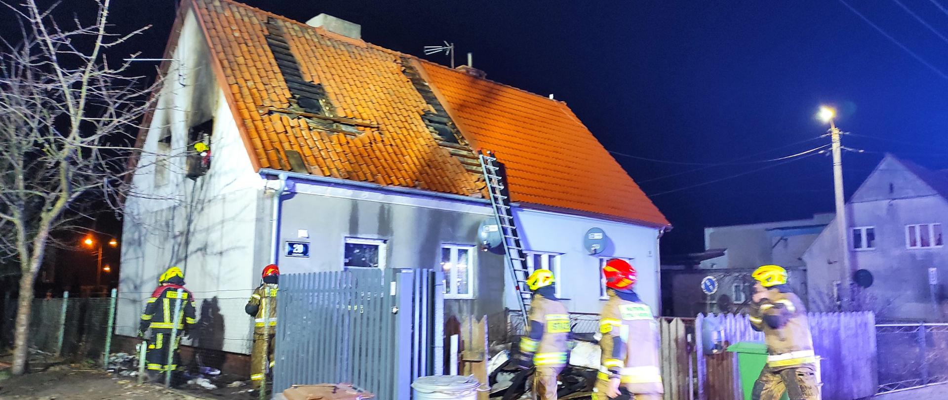 Zdjęcie zrobione nocą. Budynek oświetlony przez lampy strażackie. Dach budynku uszkodzony po pożarze. Przy budynku strażacy.