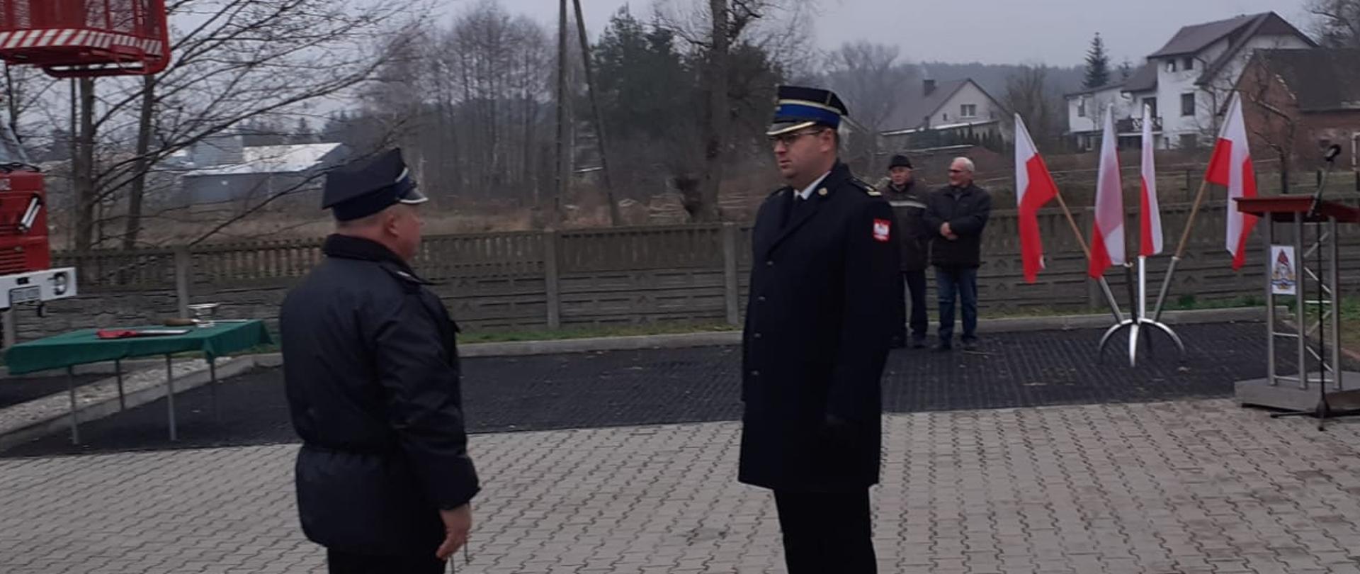 składanie meldunku przed remizą przez strażaka drugiemu strażakawowi, w tle samochód drabina oraz flagi biało czerwone 