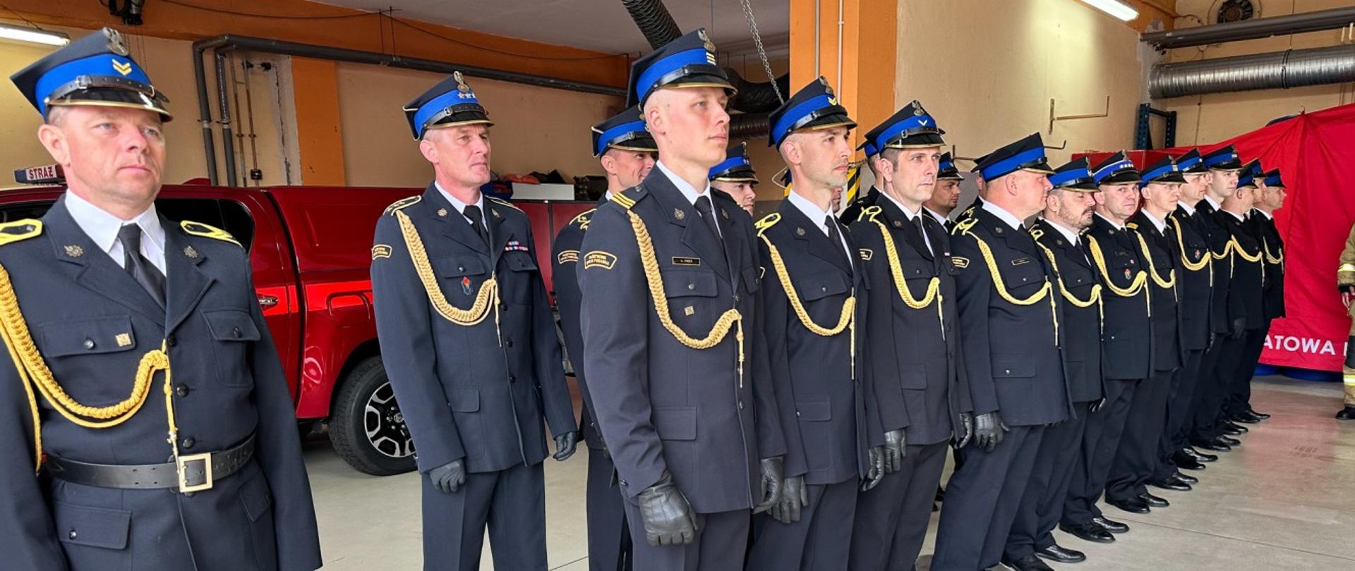Zdjęcie przedstawia funkcjonariuszy Komendy Powiatowej PSP w Wieruszowie stojących w pomieszczeniu garażowym, gdzie odbywała się uroczystość Dnia Strażaka ustawionych w dwuszeregu, w umundurowaniu wyjściowym.
