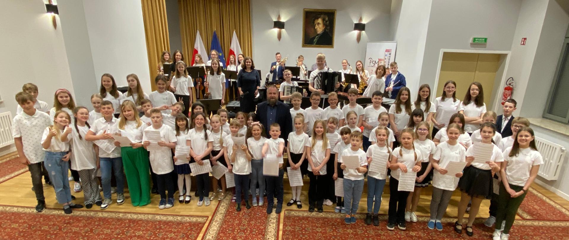 Zdjęcie przedstawia uczniów i nauczycieli szkoły muzycznej, którzy zaangażowani byli w wykonanie utworu "Zbójnicki" w dniu 25 maja 2022 r. Zdjęcie wykonano na sali koncertowej szkoły muzycznej.