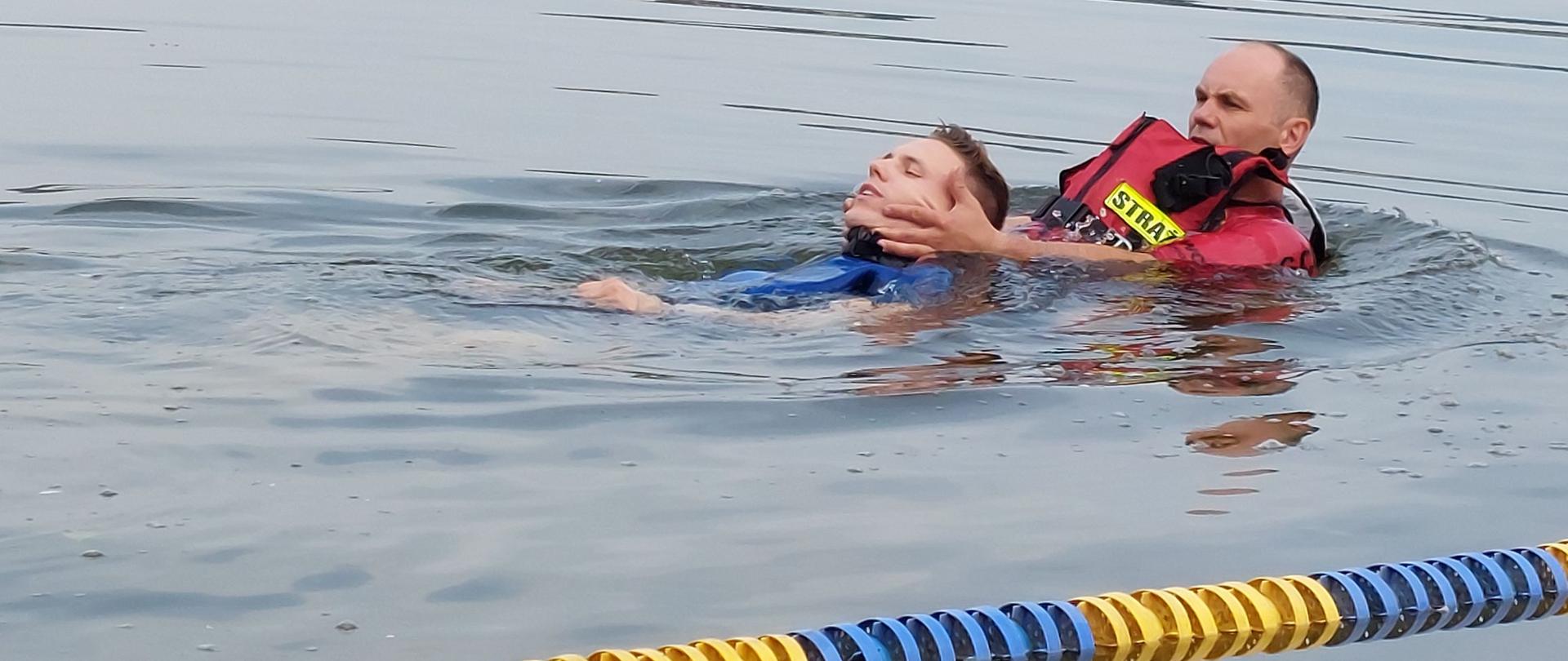 Zdjęcie przedstawia dwóch strażaków ćwiczących w wodzie ratownictwo wodne – holowanie poszkodowanego.
W tle jezioro.
