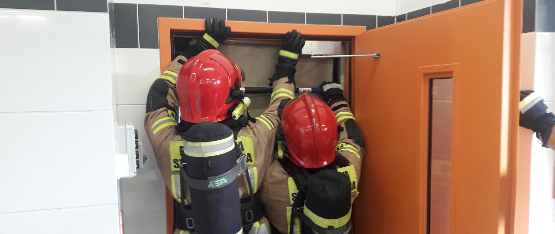 Dwóch strażaków zakłada kurtynę dymową w drzwiach. Ubrani w aparaty oddechowe.