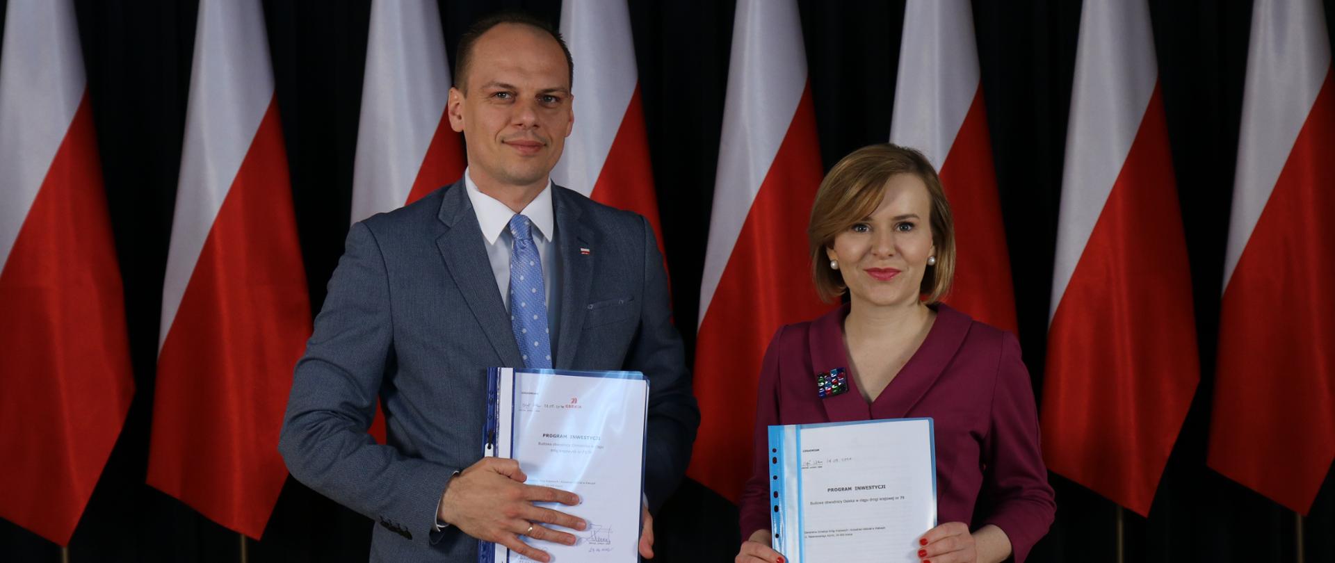 Na zdjęciu widać dwie osoby, które trzymają w rękach dokumenty. Po lewej stronie widać wysokiego mężczyznę, po prawej stronie kobietę. W ich tle stoją biało-czerwone flagi.