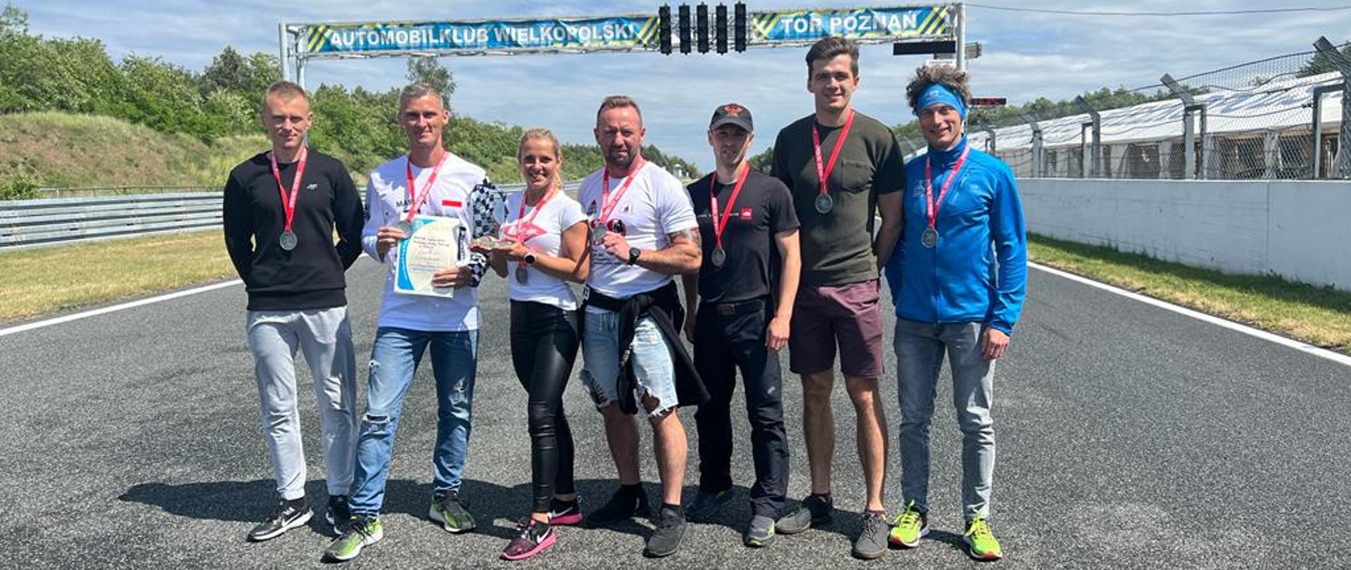 Zdjęcie przestawia 7 osób stojących w rzędzie na asfaltowej nawierzchni z medalami zawieszonymi na szyi. W tle bramownica z napisem Automobilklub Wielkopolski Tor Poznań.