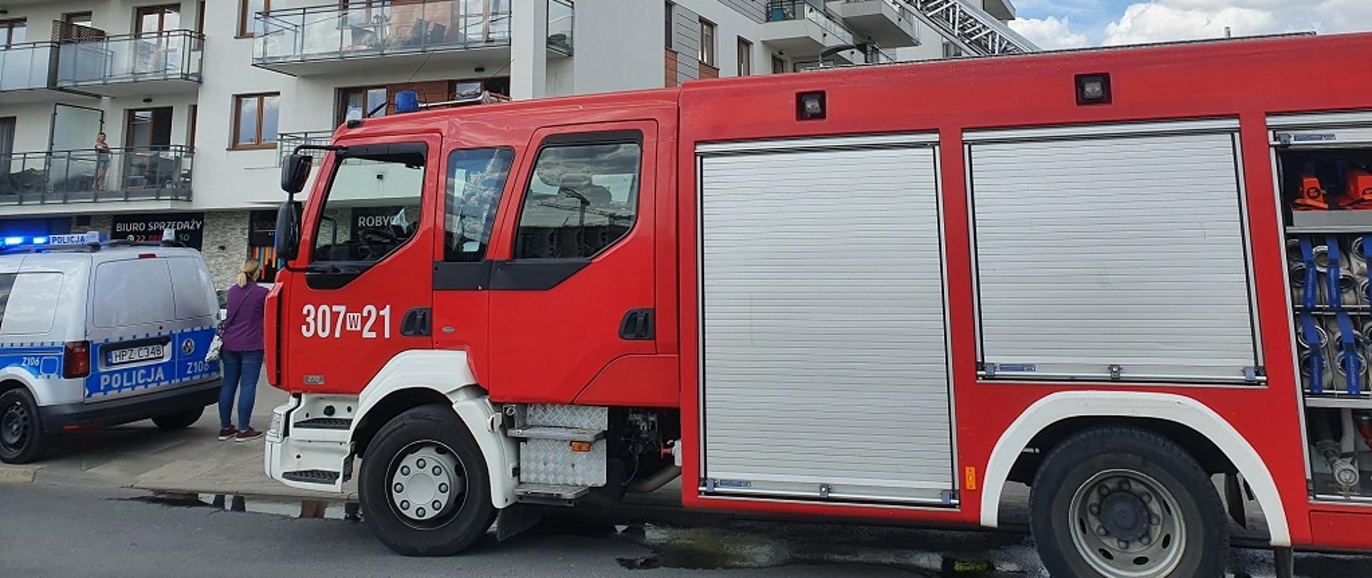 Na pierwszym planie zdjęcia widać bok średniego samochodu ratowniczo-gaśniczego w czerwonym kolorze z białym oznakowaniem operacyjnym: 307 W 21 oraz szarymi skrytkami sprzętowymi. Na drugim palnie zdjęcia widoczny jest budynek mieszkalny wielorodzinny