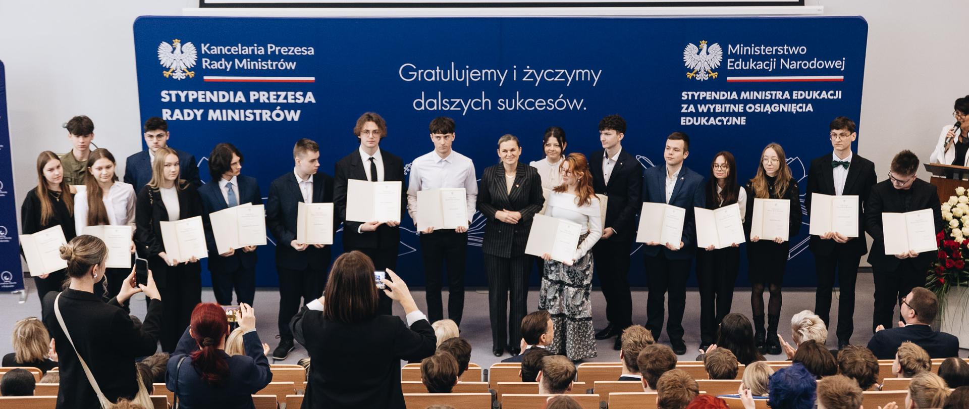 Druga grupa stypendystów pozuje na scenie z Barbarą Nowacką na tle niebieskiej ścianki informacyjnej. Minister stoi w środku, uśmiechając się i trzymając splecione dłonie. Przed sceną widać siedzącą publiczność oraz osoby robiące zdjęcia telefonami.