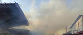 Teren tartaku, widoczne duże zadymienie, z prawej strony zdjęcia budynek mieszkalny, z lewej wiaty w których znajdowały się maszyny do przecierania drewna, na wprost zniszczona przez ogień hala oraz strażacy prowadzący działania.