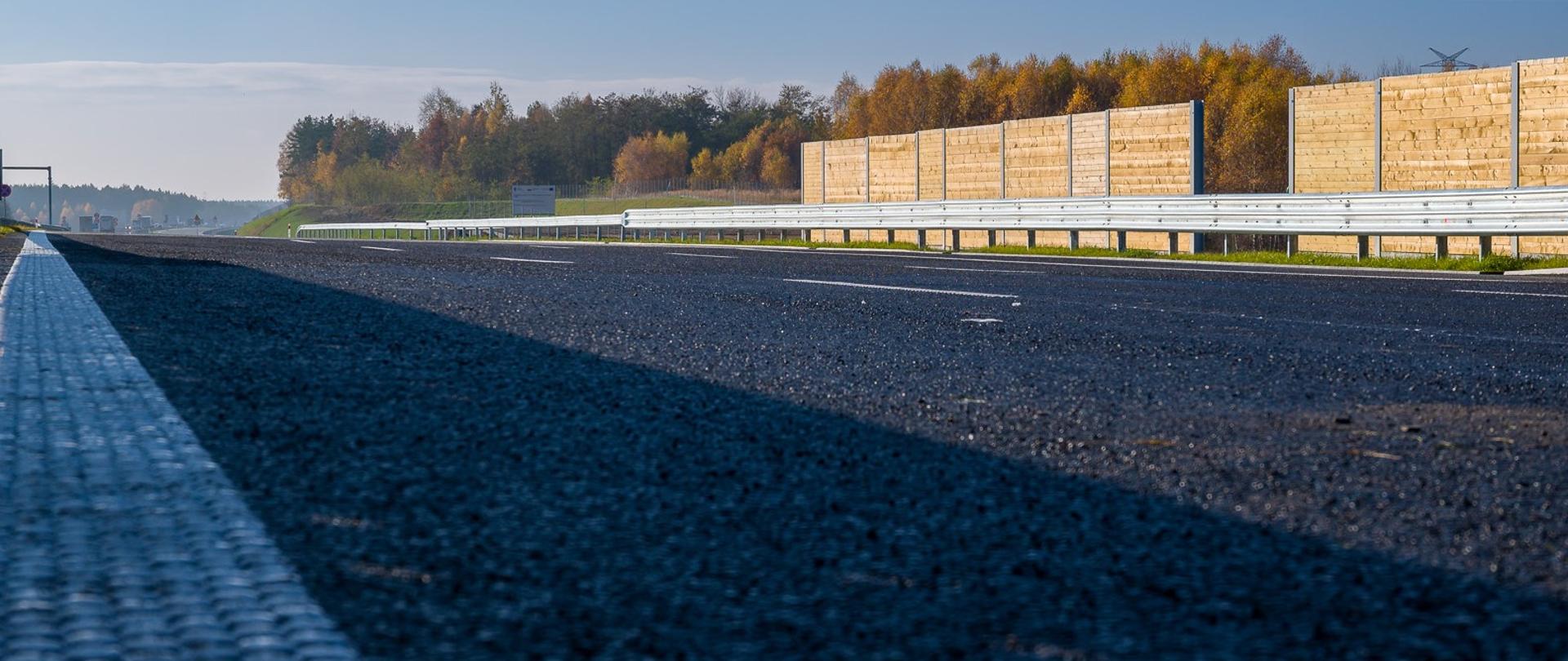Słoneczny dzień. Zbliżenie na jezdnię autostrady A1. W tle widoczne bariery, a za nimi drzewa.