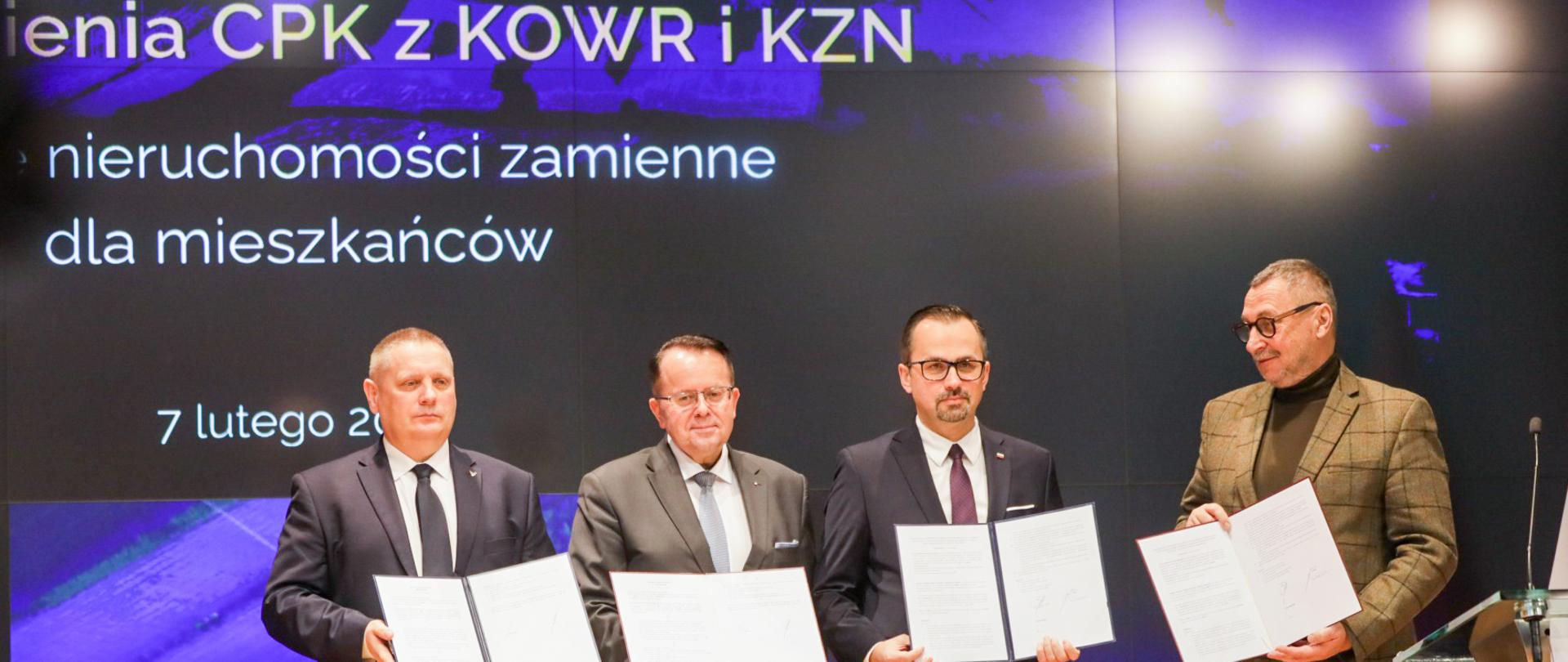 Cztery osoby stoją na scenie i trzymają w rękach podpisane dokumenty.