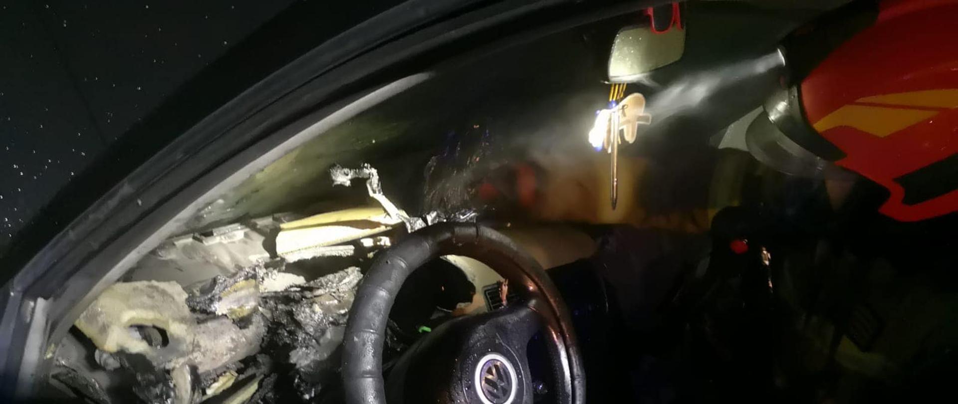 Na fotografii znajduje się wnętrze spalonego pojazdu. Zdjęcie zostało wykonane od strony szyby kierowcy. Widać całkowicie stopioną deskę rozdzielczą, która od ognia zrobiła się czarna. Po prawej górnej stronie znajduje się strażak w czerwonym hełmie, który zagląda do środka pojazdu. Ma włączoną latarkę i oświetla kierownicę - zdjęcie wykonano w nocy.