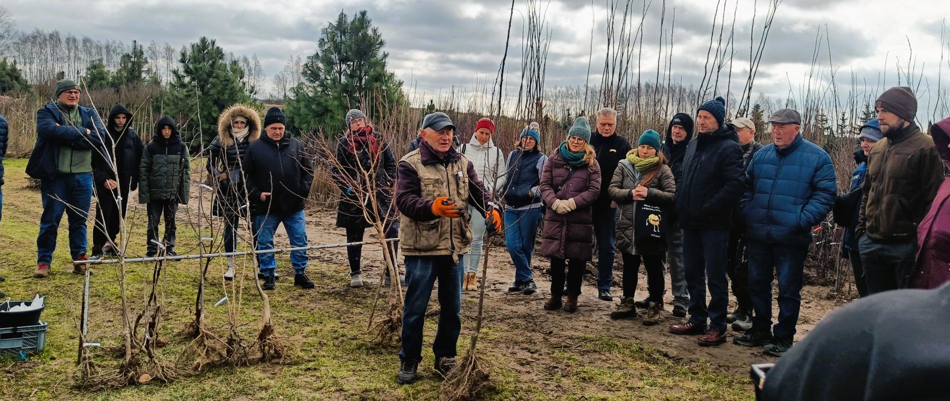 Ludzie w kurtkach stoją wokół osoby trzymającej drzewko bez liści