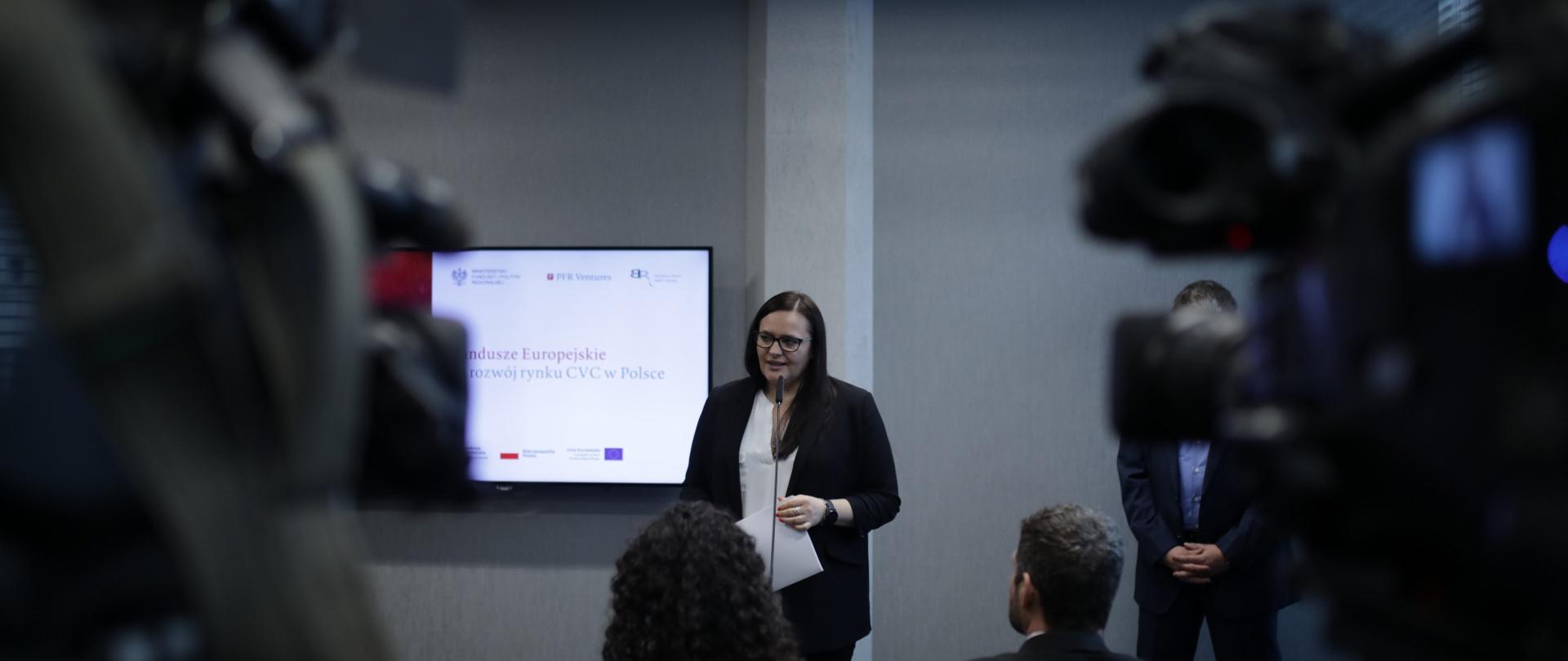 Minister Jarosińska-Jedynak stoi przy mikrofonie, do którego mówi, przed nią stoją kamery