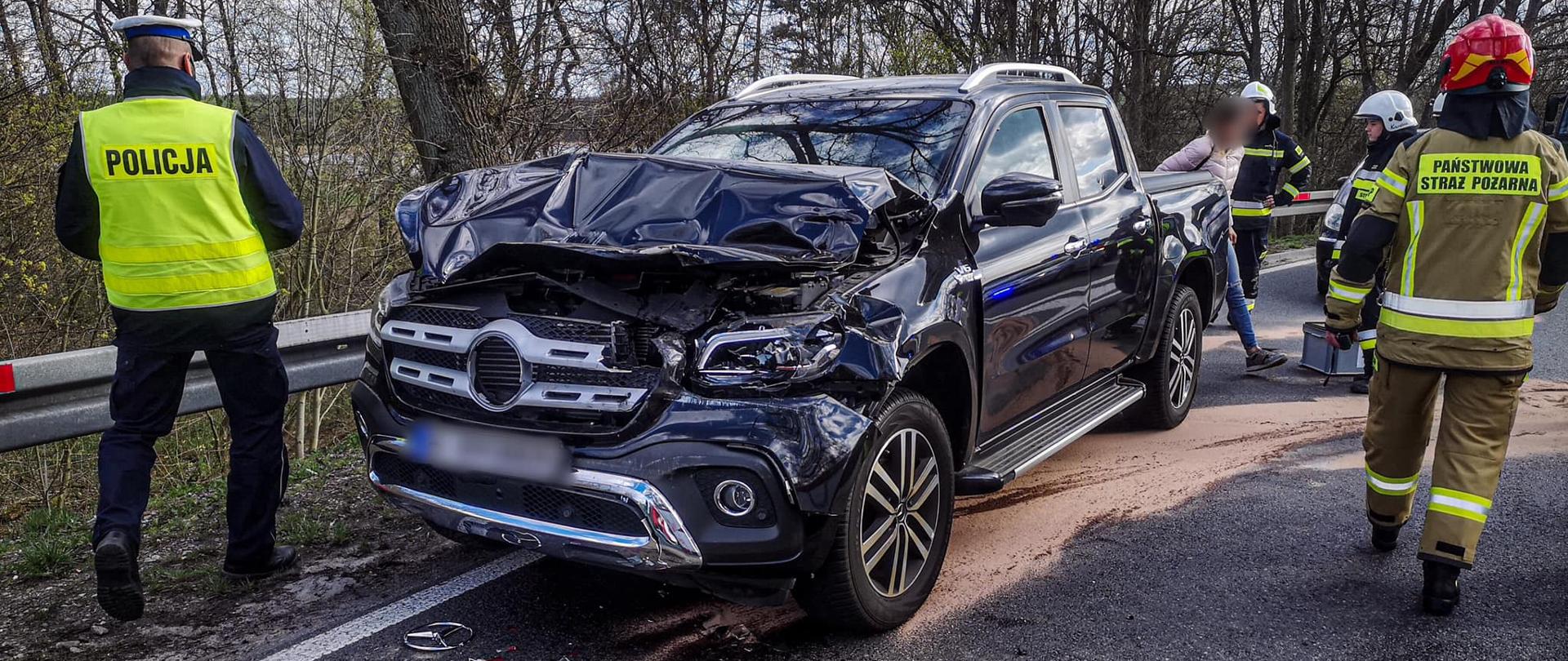Na fotografii znajduje się czarny samochód osobowy, który ma uszkodzony przód. Dookoła pojazdu znajdują się strażacy oraz policjanci. Na asfalcie znajdują się rozwalone elementy rozbitego pojazdu. W tle znajdują się drzewa. Zdjęcie wykonano w porze dziennej.