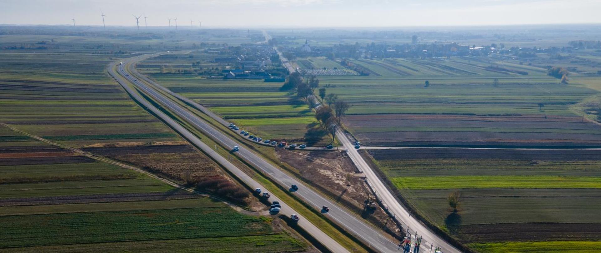 Na zdjęciu widać obwodnicę Dzwoli z lotu ptaka. Na jezdni znajdują się ludzie w kamizelkach oraz samochody. Po obu stronach widać pola, a w tle wiatraki.