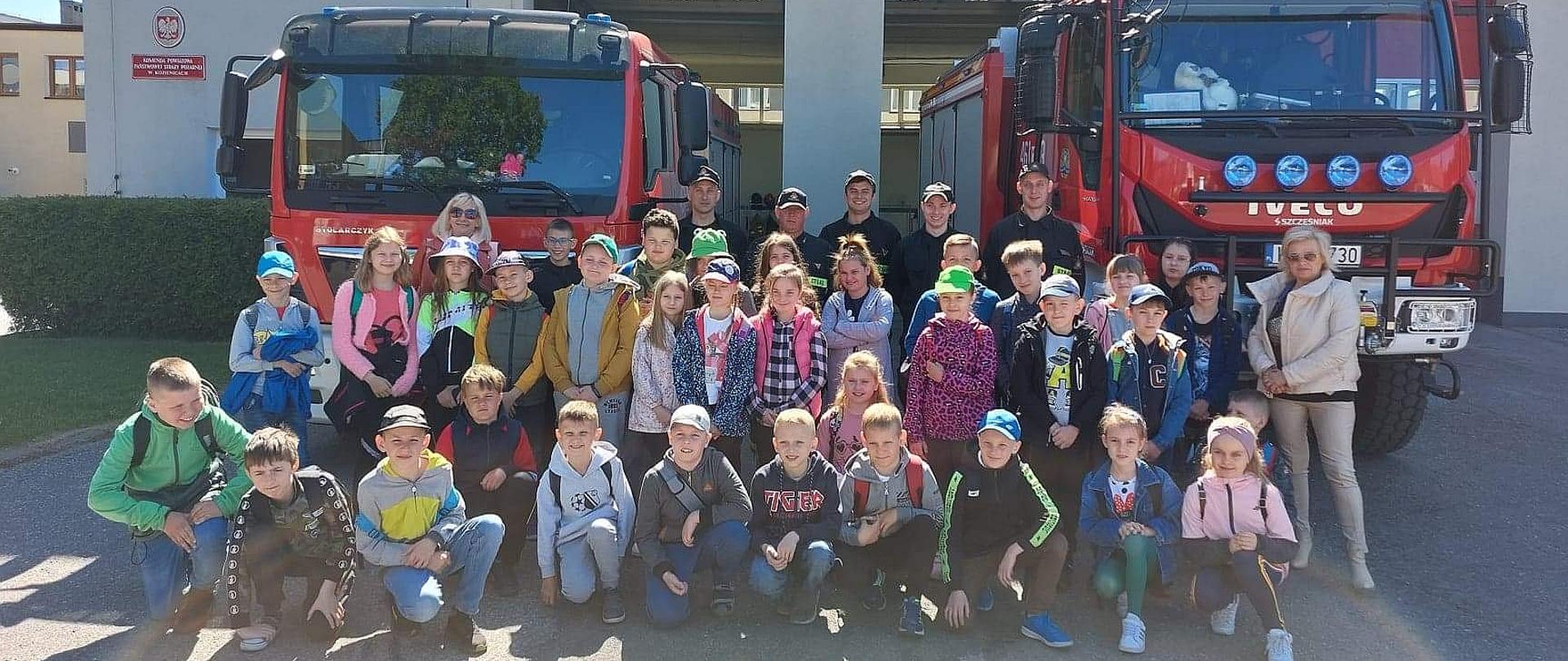 grupa uczniów ze strażakami pozują do zdjęcia grupowego na placu strażnicy w Kozienicach na tle samochodów strażackich wystawionych przed bramy garażowe
