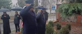 Na tle drzew i budynków stoi dwóch funkcjonariuszy Służby Więziennej i Państwowej Straży Pożarnej. Maja na sobie umundurowanie wyjściowe zimowe. Oddają honor, poprzez salutowanie, Pomnikowi Działaczy Solidarności, po uprzednim złożeniu wiązanki kwiatów. Za nimi pod parasolkami znajdują się inni uczestnicy wydarzenia. 