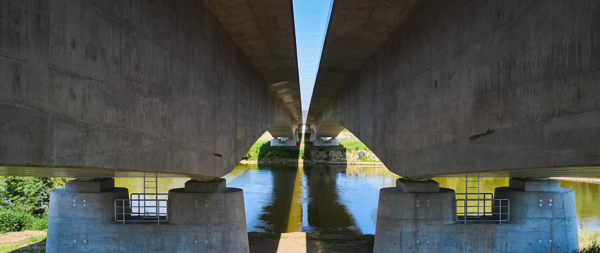 Widok z dołu na most na rzece San w ciągu S19. Widok z brzegu na rzekę, podpory i przęsła. Patrząc w górę widać przestrzeń między jezdniami zabezpieczoną siatkami ochronnymi. 