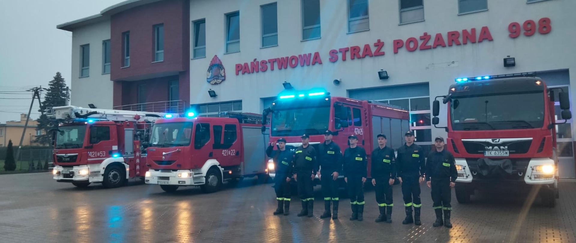 Strażacy na zbiórce przed jednostką na tle samochodów pożarniczych. Pojazdy mają włączone sygnały świetlne.