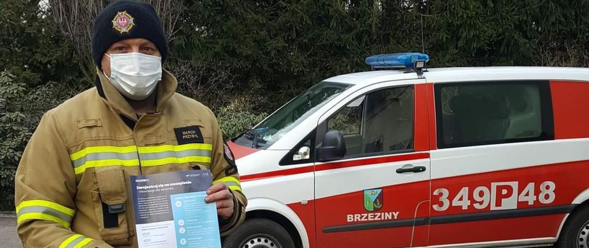 Na zdjęciu widać strażaka, który trzyma niebieską ulotkę informacyjną o szczepieniach przeciwko COVID. Za strażakiem jest samochód strażacki w kolorze czerwonym z umieszczoną na dachu sygnalizacją ostrzegawczą. Na boku widać numery operacyjne. Strażak stoi na kostce brukowej. Za samochodem strażackim widać zielone drzewa iglaste.

