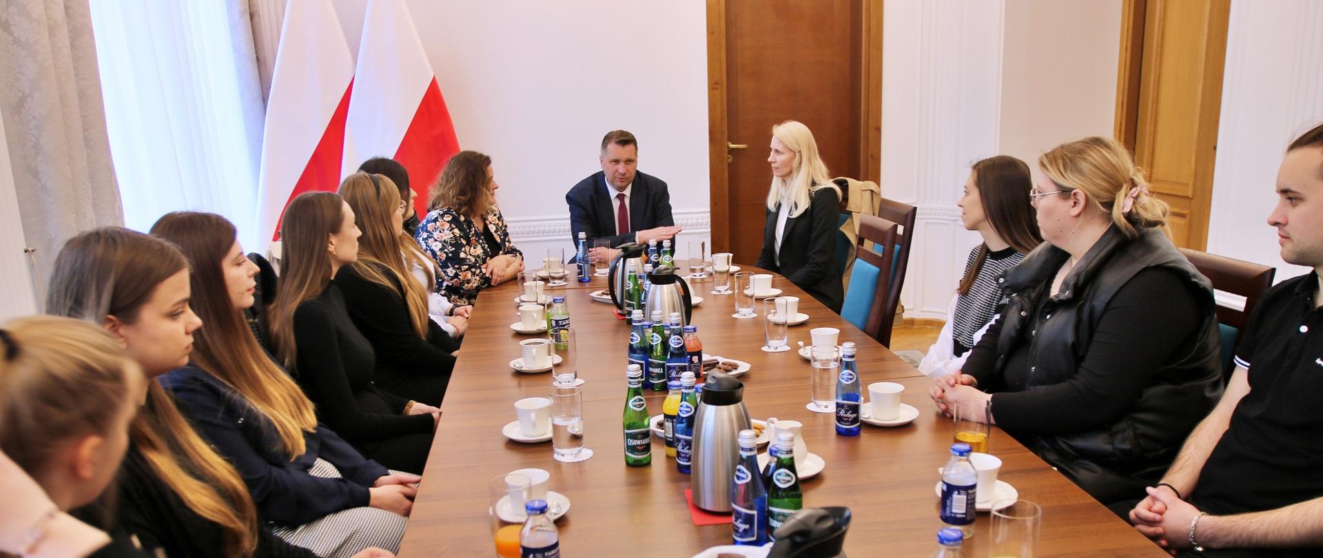 Po obu stronach długiego prostokątnego stołu siedzi kilkanaście młodych osób, u szczytu stołu minister Czarnek, za nim dwie polskie flagi i biała ściana z drewnianymi drzwiami, po lewej zasłonięte firankami okna.