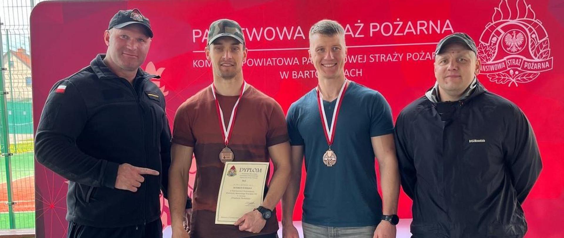 Czterech mężczyzn stoi na tle czerwonego banneru Państwowej Straży Pożarnej. Dwóch z nich ma medale oraz trzyma w dłoni dyplomy. Na ziemi leżą nagrody.