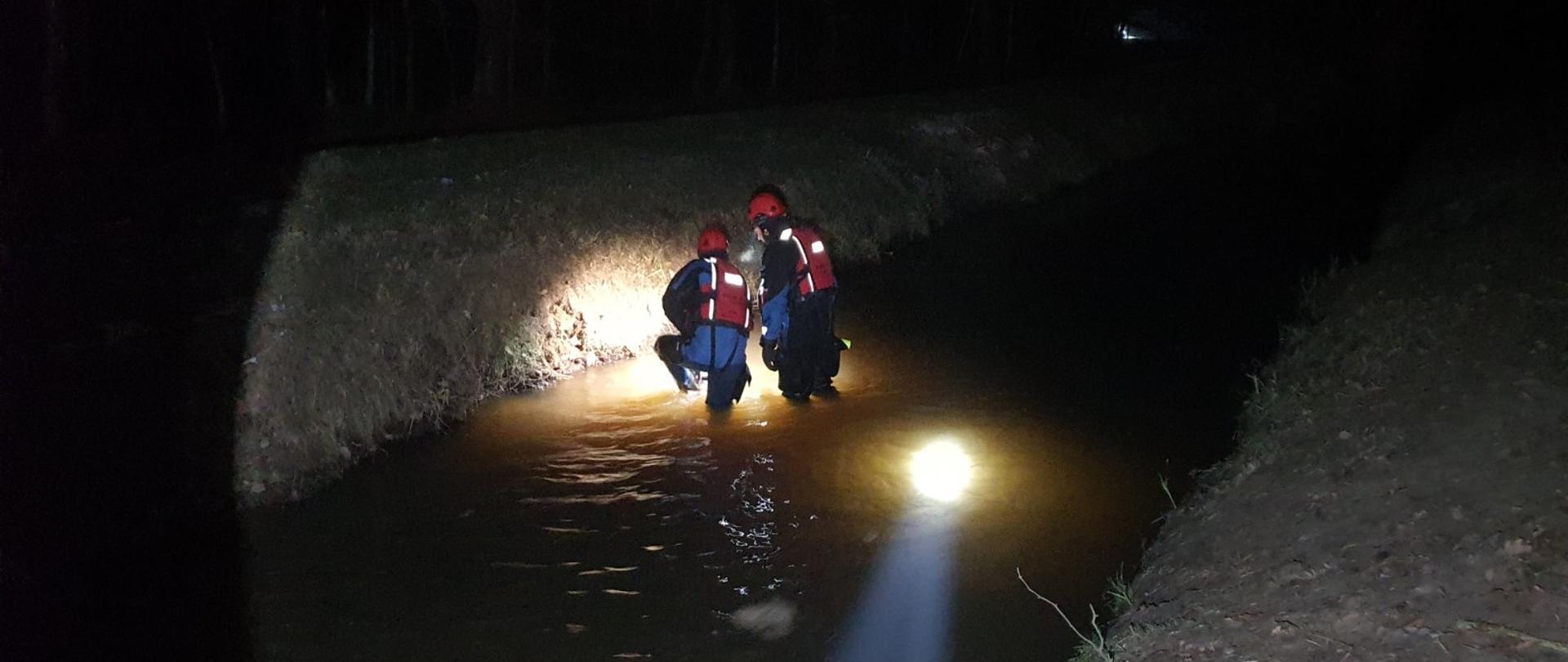 Kanał wodny nocą. W kanale dwóch ratowników ubranych w skafandry do pracy w wodzie. Ratownicy penetrują kanał. 