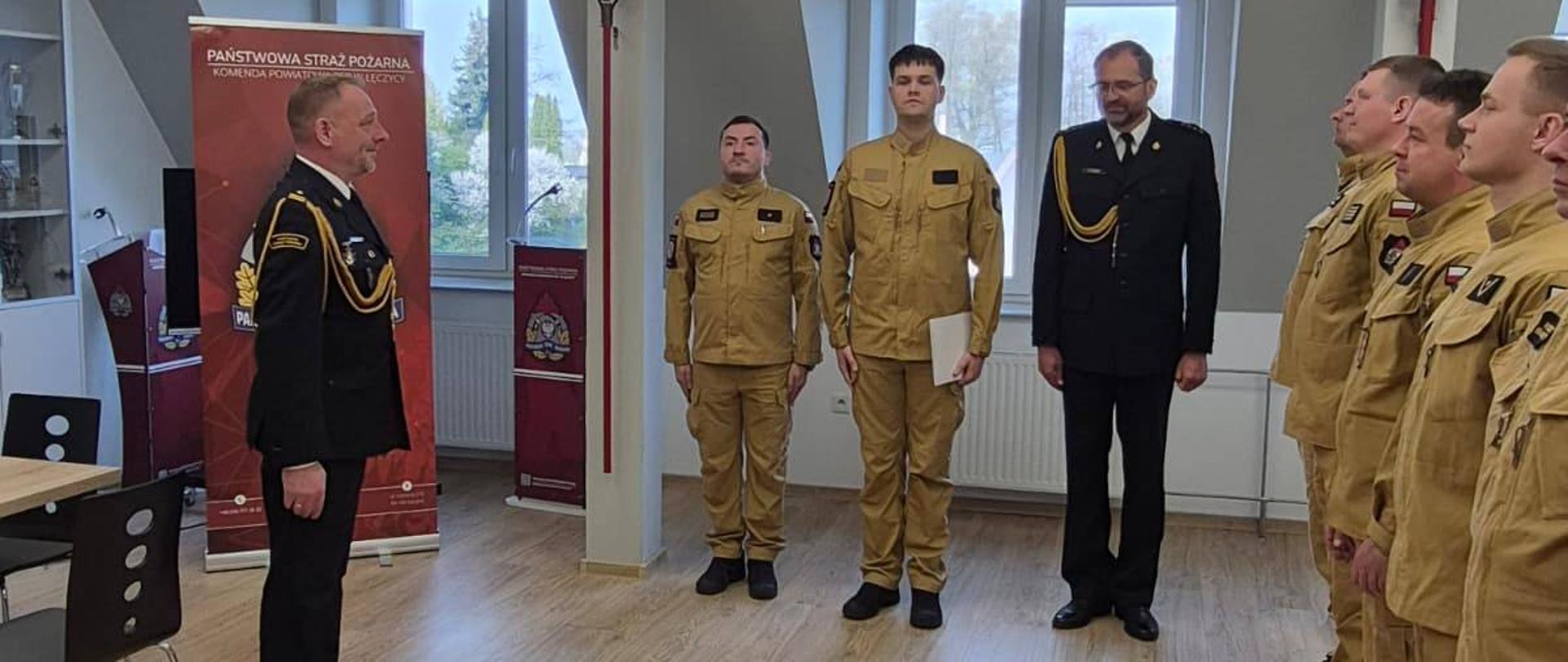 Na zdjęciu widoczna jest grupa mężczyzn w mundurach strażackich. Po prawej stronie ustawieni są w szeregu funkcjonariusze w piaskowych uniformach, stojący na baczność. Naprzeciw nich stoi mężczyzna w galowym mundurze. W tle widać okna oraz dwóch funkcjonariuszy w piaskowym mundurze i jednego mężczyznę w galowym mundurze. Po lewej stronie widoczne są gabloty oraz baner z napisem „Państwowa Straż Pożarna”.