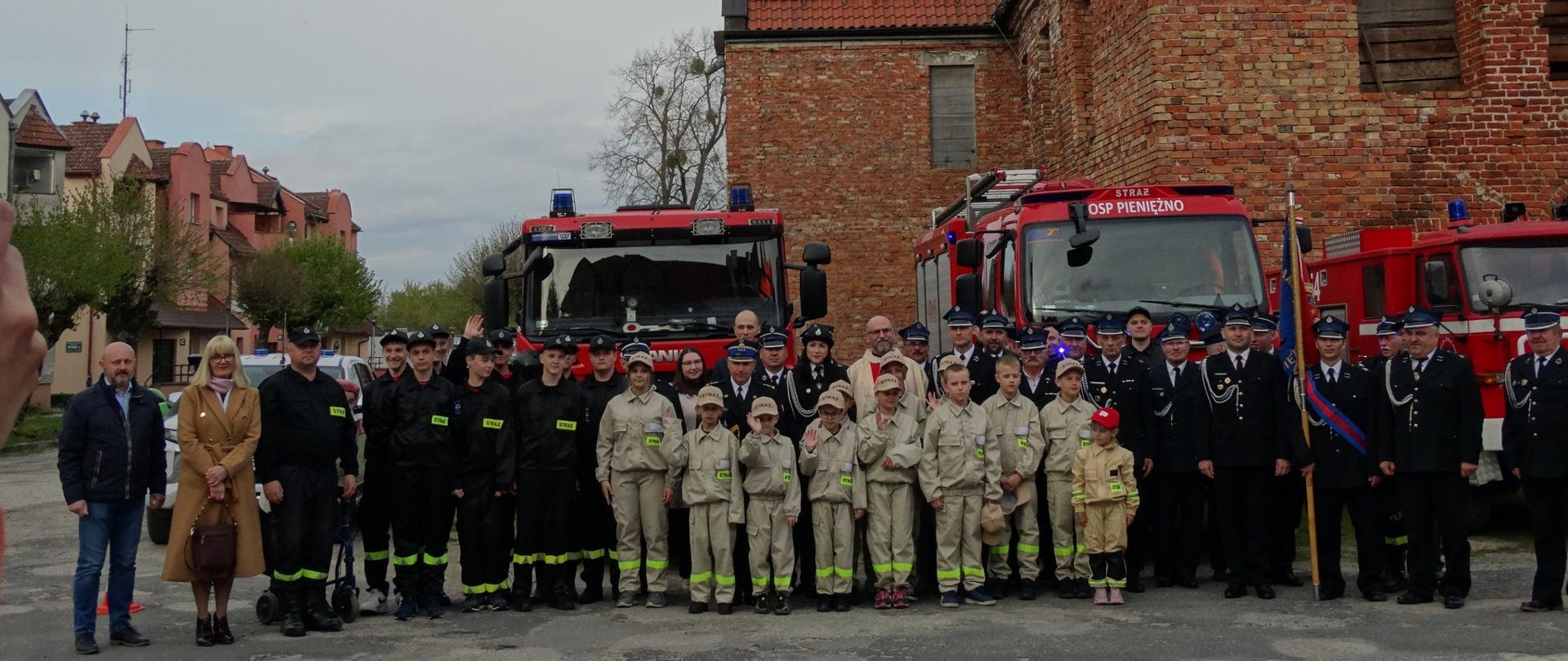 Na zdjęciu na tle zabytkowego budynku z cegły stoi duża grupa strażaków w mundurach wyjściowych i dzieci z młodzieżowej drużyny pożarniczej, za nimi samochody strażackie.