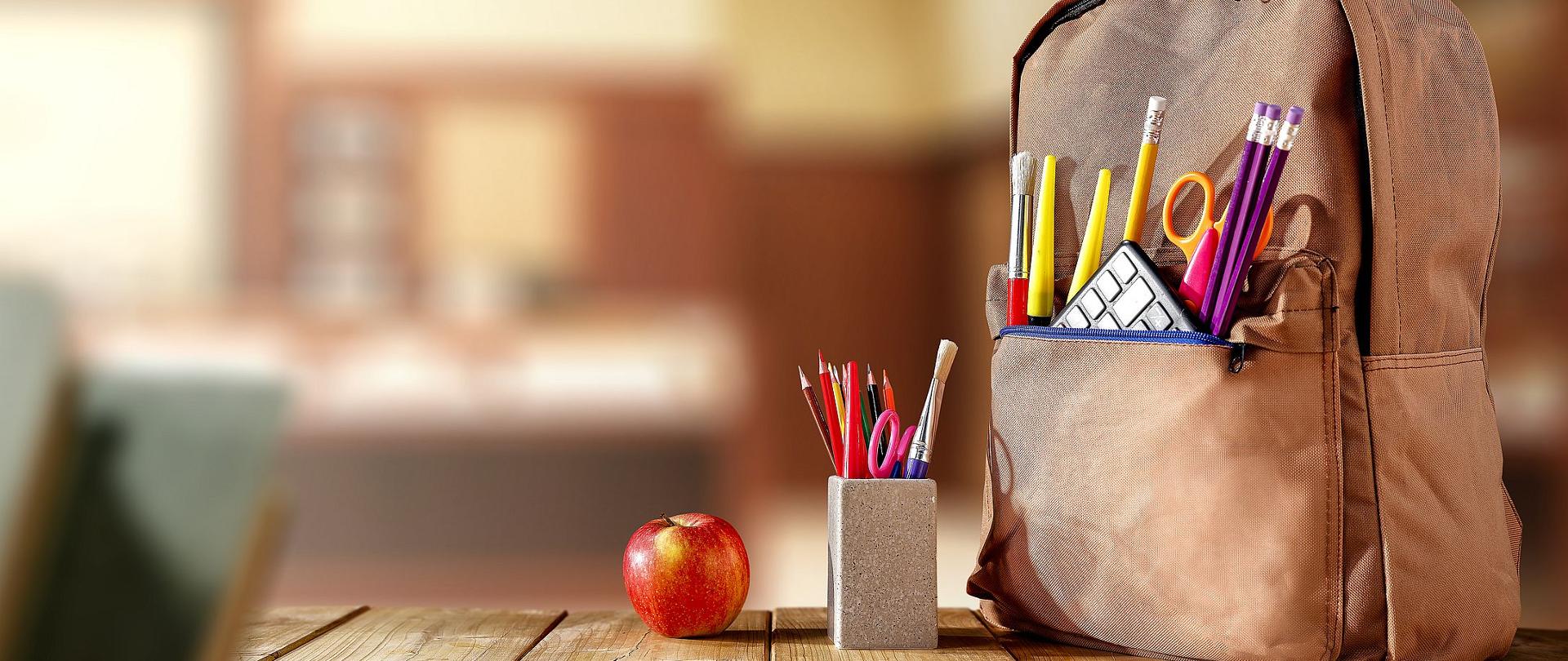 Zdjęcie przedstawia położony na szkolnym biurku plecak, pojemnik na przybory szkolne oraz jabłko. W tle rozmyte tło sali szkolnej.