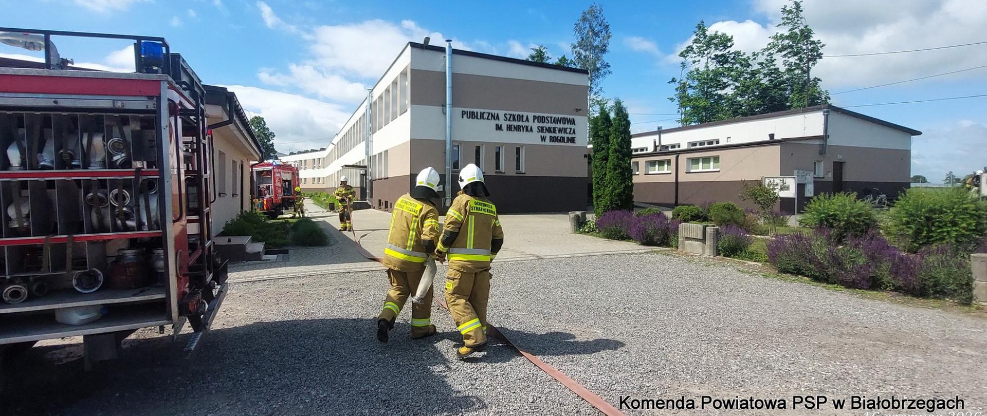 Trzej strażacy w pełnym umundurowaniu bojowym i białych hełmach rozwijają linię gaśniczą z węża pożarniczego na żwirowym placu przed budynkiem Publicznej Szkoły Podstawowej im. Henryka Sienkiewicza w Rogolinie. Po lewej stronie widoczny jest tył wozu strażackiego z wyposażeniem, a w tle drugi czerwony samochód ratowniczo-gaśniczy.