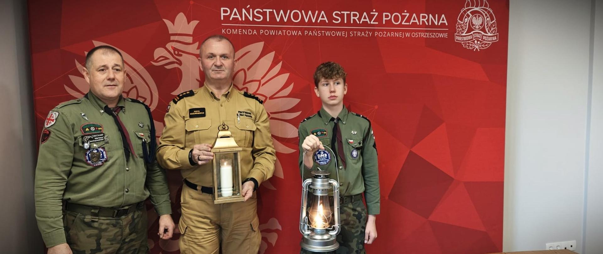 Harcerze przekazują światło komendantowi straży pożarnej. Osoby pozują na tle czerwonego baneru firmowego.