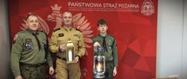 Harcerze przekazują światło komendantowi straży pożarnej. Osoby pozują na tle czerwonego baneru firmowego.