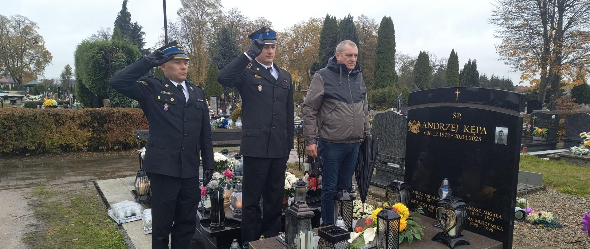 Na zdjęciu przed grobem stoi dwóch strażaków salutujących i jeden emeryt strażacki