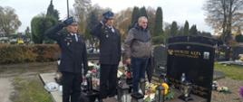 Na zdjęciu przed grobem stoi dwóch strażaków salutujących i jeden emeryt strażacki