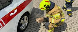 Na zdjęciu strażak w hełmie podkłada drewniany klocek pod koło samochodu pożarniczego