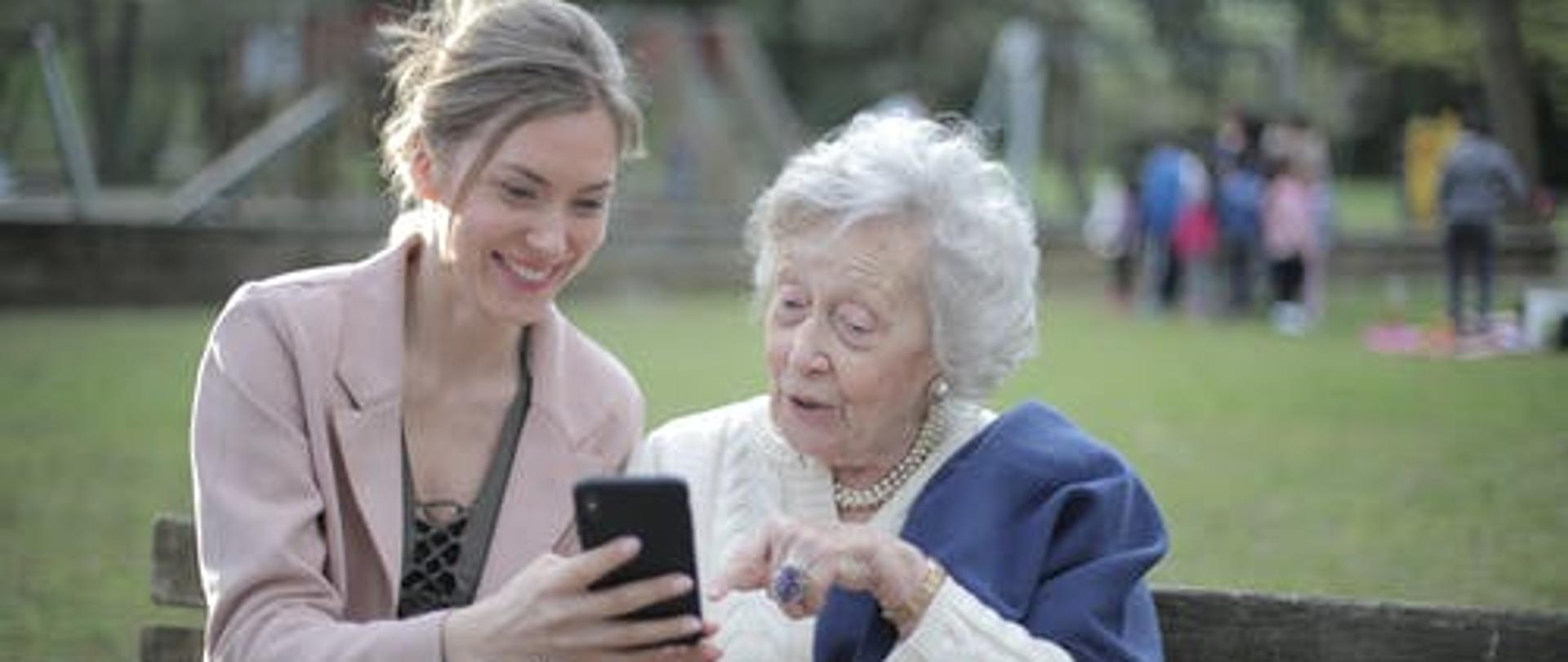 Park. Na ławce starsza pani. Obok niej siedzi dziewczyna pokazując coś na ekranie telefonu. W tle drzewa i zarysy innych postaci.