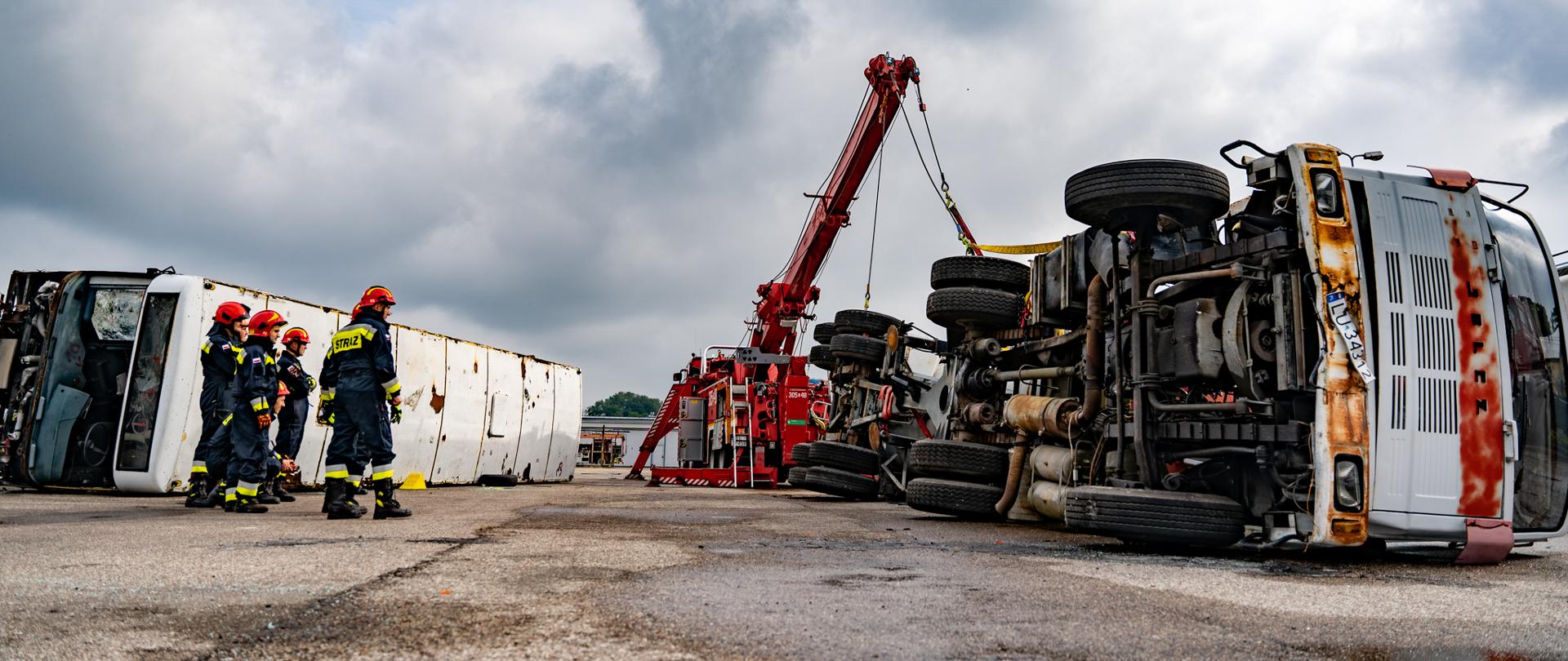 Grupa strażaków w kombinezonach obok przewróconego samochodu ciężarowego