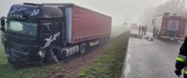 Pora dzienna, duże zamglenie. Po lewej stronie kadru widać ciężarówkę z naczepą stojącą na polu obok drogi. po prawej stoi samochód pożarniczy, koło niego trzech ratowników. W oddali widać parawan ratowniczy i kolejny pojazd pożarniczy.